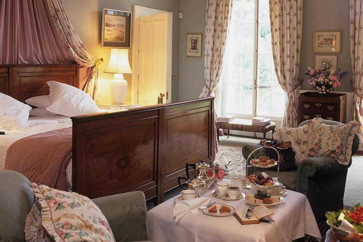 A room with a bed, a bedside table with a lamp, a small table with tea and pastries, and a chair.