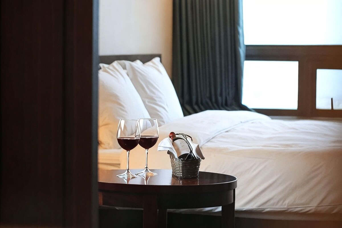 A hotel room with a bed, a table with two glasses of red wine, and a basket with a napkin.