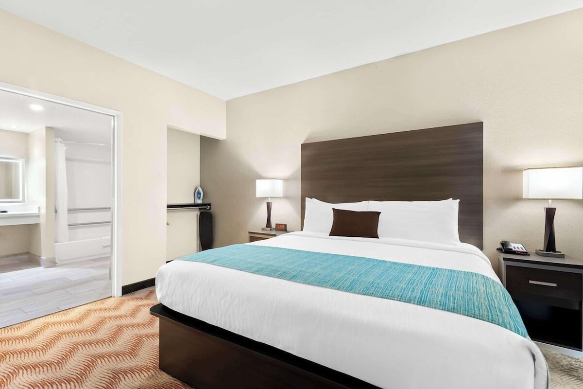 A hotel room with a large bed, a nightstand, a lamp, a mirror, and a door to a bathroom.