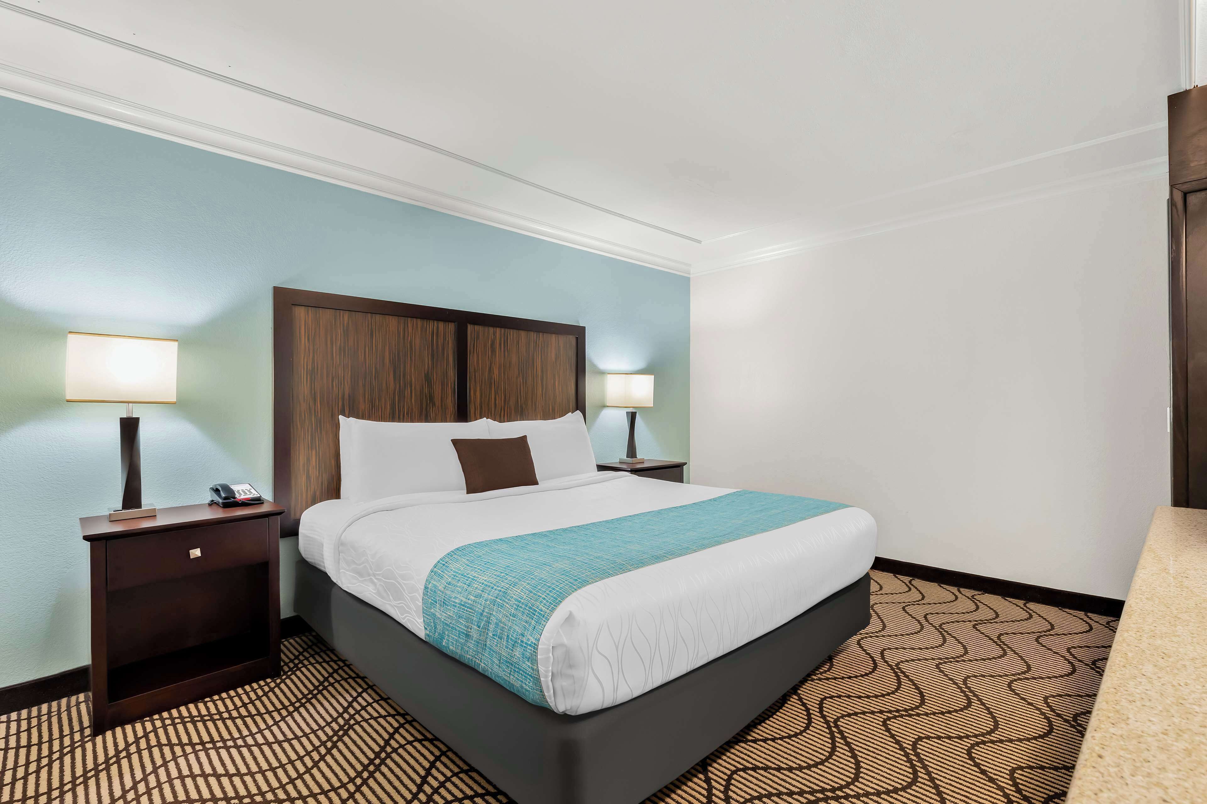 A hotel room with a bed, two bedside tables, a nightstand, a lamp, and a patterned carpet.