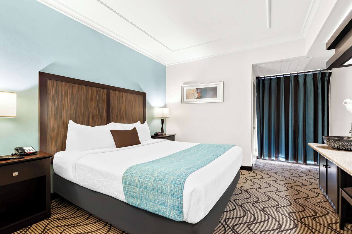 A hotel room with a large bed, a desk with a bowl, a nightstand with a phone, a picture on the wall, and a window with curtains.