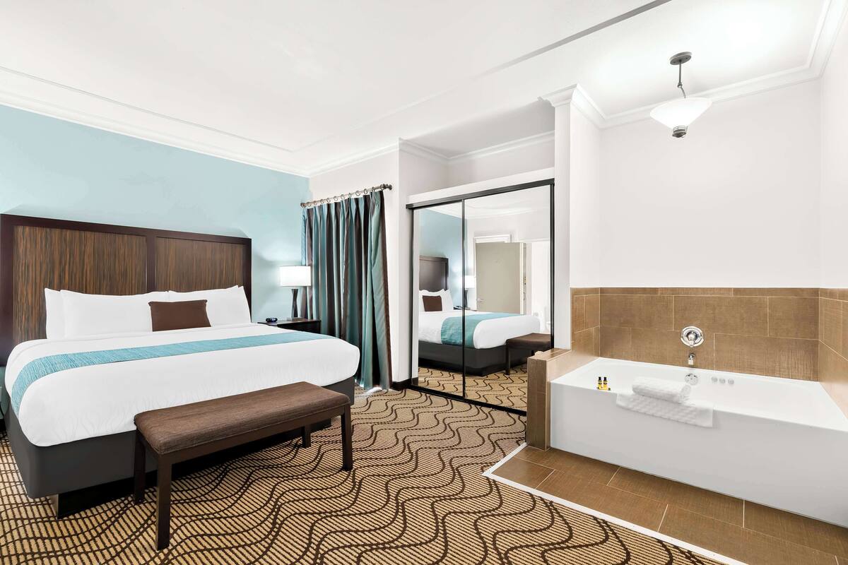 A hotel room with a large bed, a bench, a bathroom with a bathtub, and a mirror.