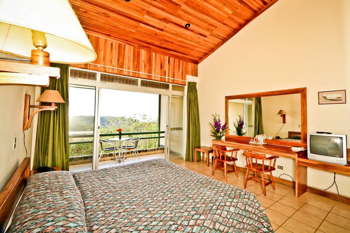 Un dormitorio con cama, escritorio con televisor, balcón con silla y ventana grande.