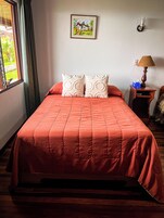 Una cama con una colcha roja, dos almohadas decorativas, un cuadro enmarcado en la pared, una mesita de noche con una lámpara y una ventana con cortinas.