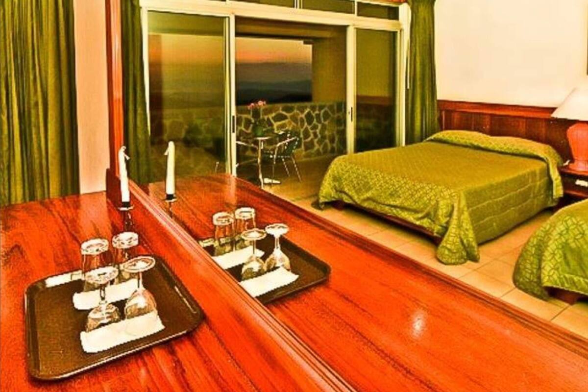 Habitación de hotel con techo de madera, cama con ropa de cama verde, mesa de comedor con vasos y una bandeja, y puerta corredera de vidrio que da a un área exterior.