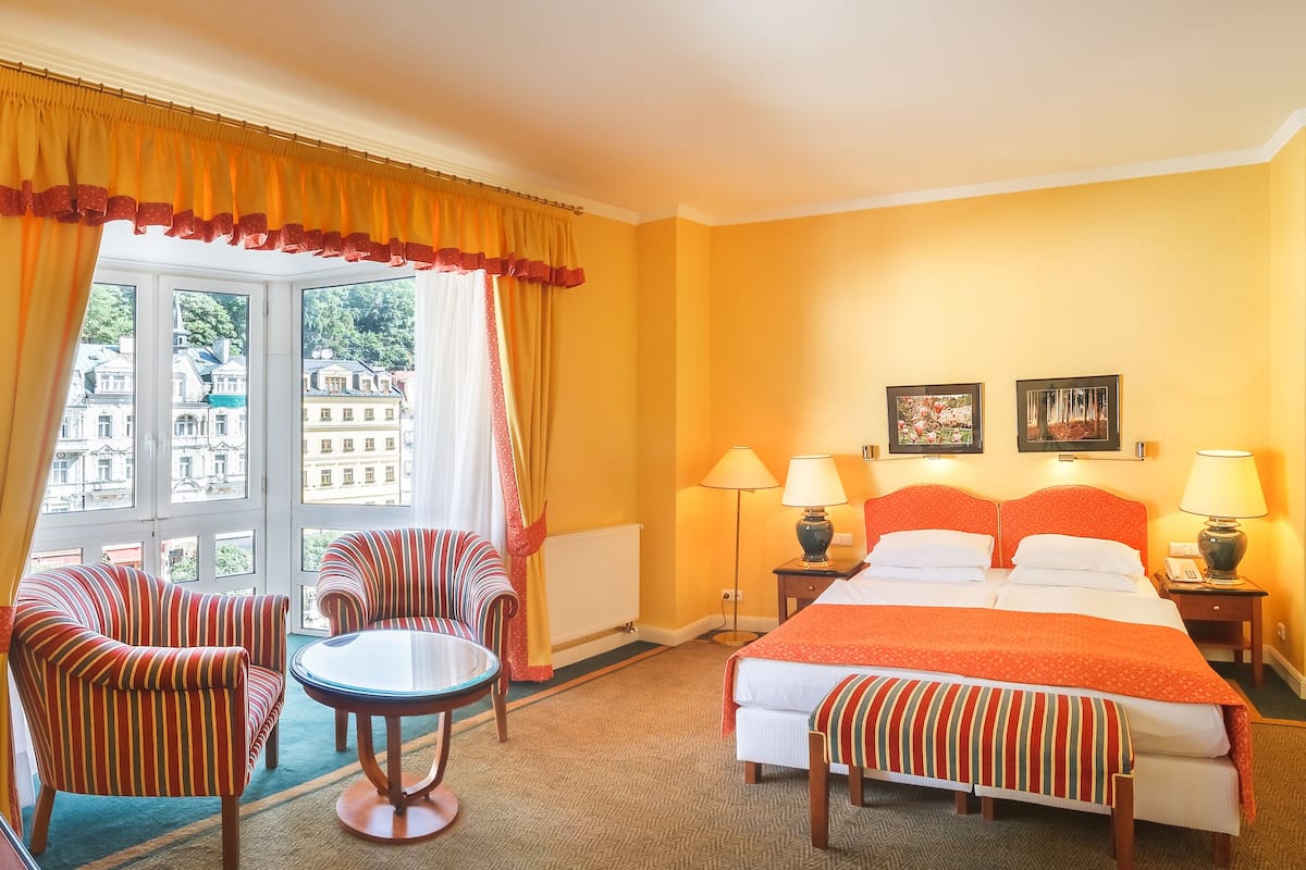 A hotel room with a bed, two chairs, a small table, and a view of buildings through large windows.