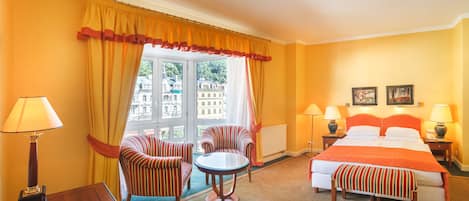 A hotel room with a bed, two chairs, a small table, and a view of buildings through large windows.