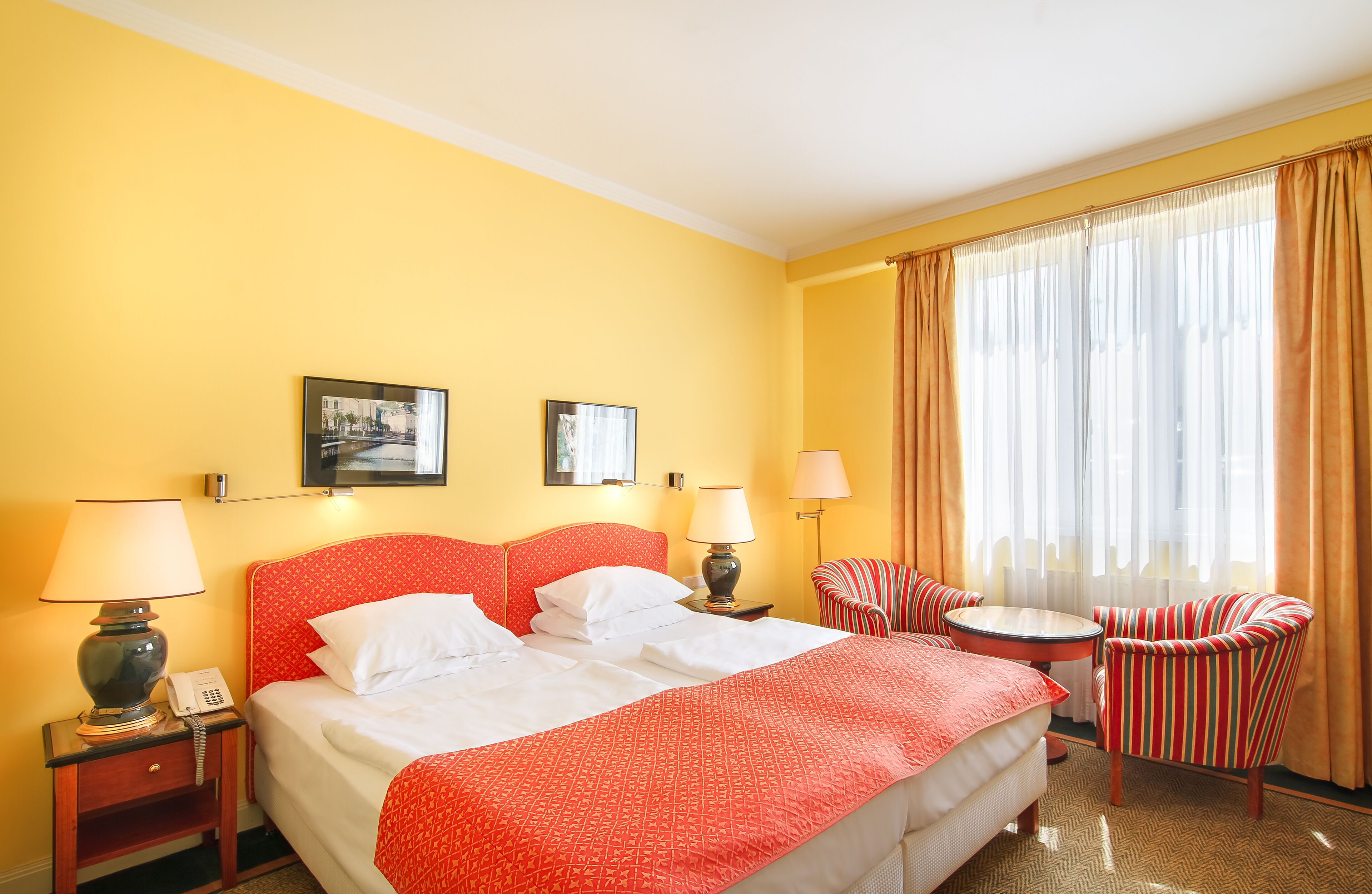 A hotel room with a bed, two bedside tables with lamps, a chair, a small round table, and a window with curtains.