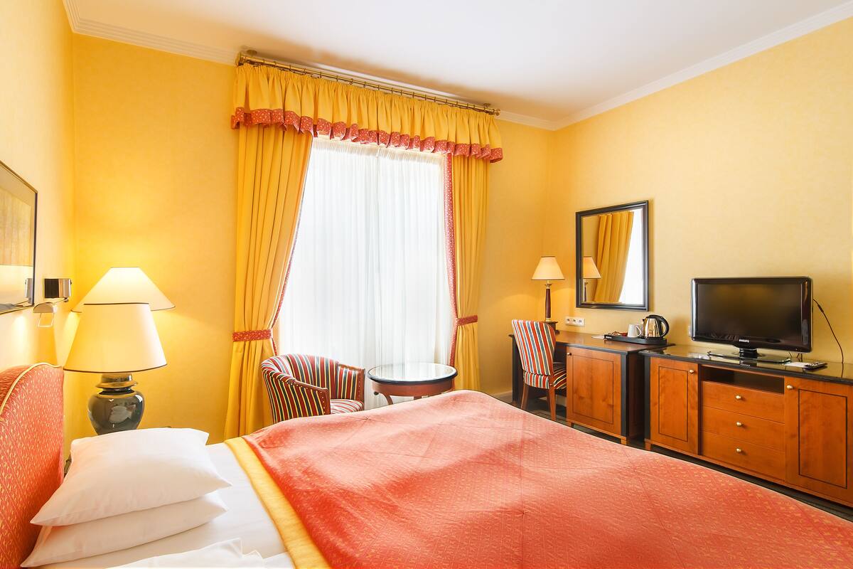 A hotel room with a bed, two chairs, a small table, a television, and a window with curtains.
