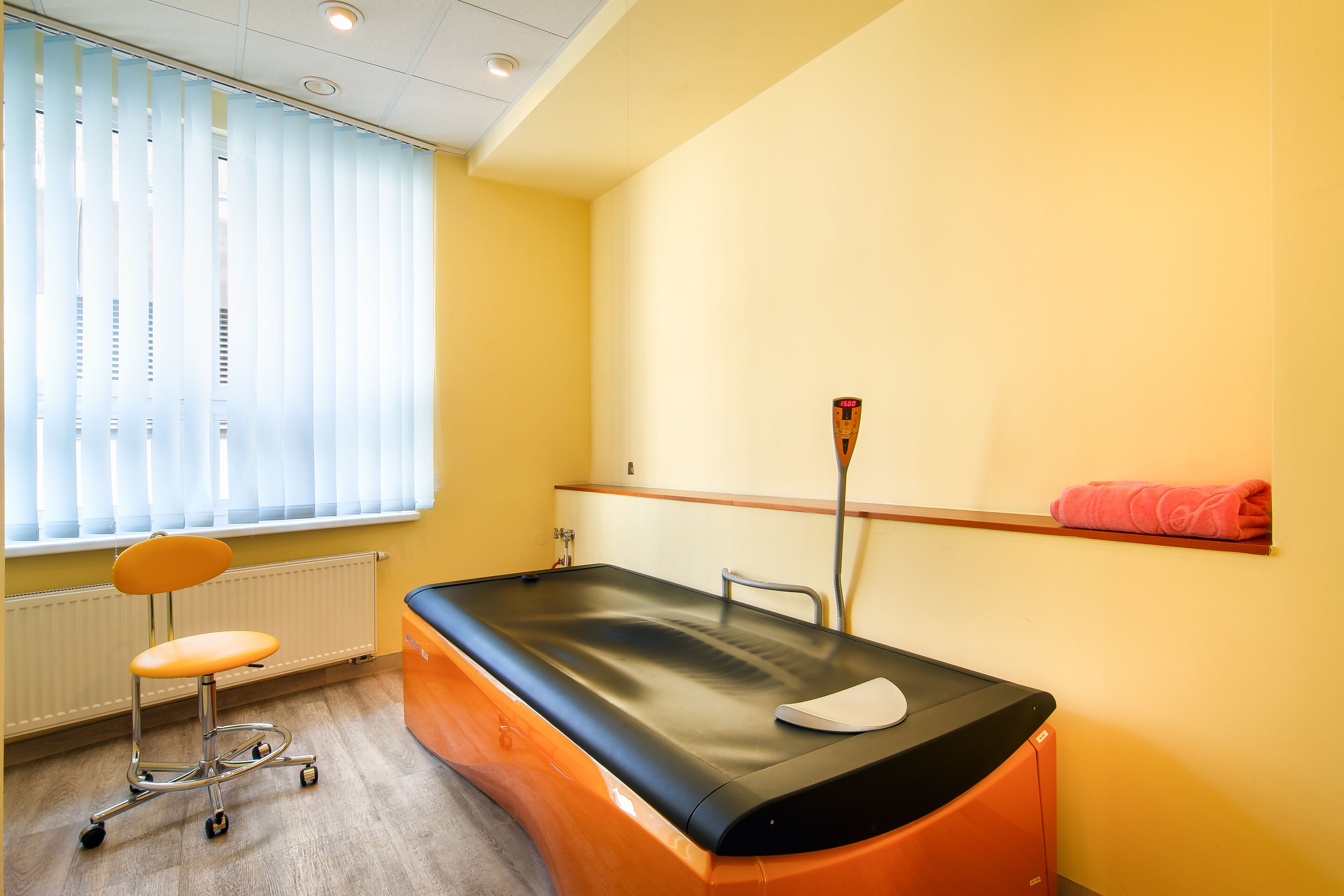 A medical examination room with a treatment table, a patient chair, a window with blinds, and a wall-mounted shelf with a rolled-up towel.
