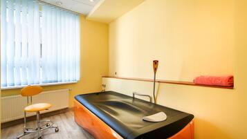 A medical examination room with a treatment table, a patient chair, a window with blinds, and a wall-mounted shelf with a rolled-up towel.