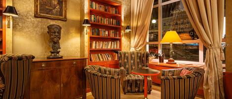 Un acogedor salón de lectura con un escritorio de madera, sillones a rayas, una estantería y un amplio ventanal con cortinas.