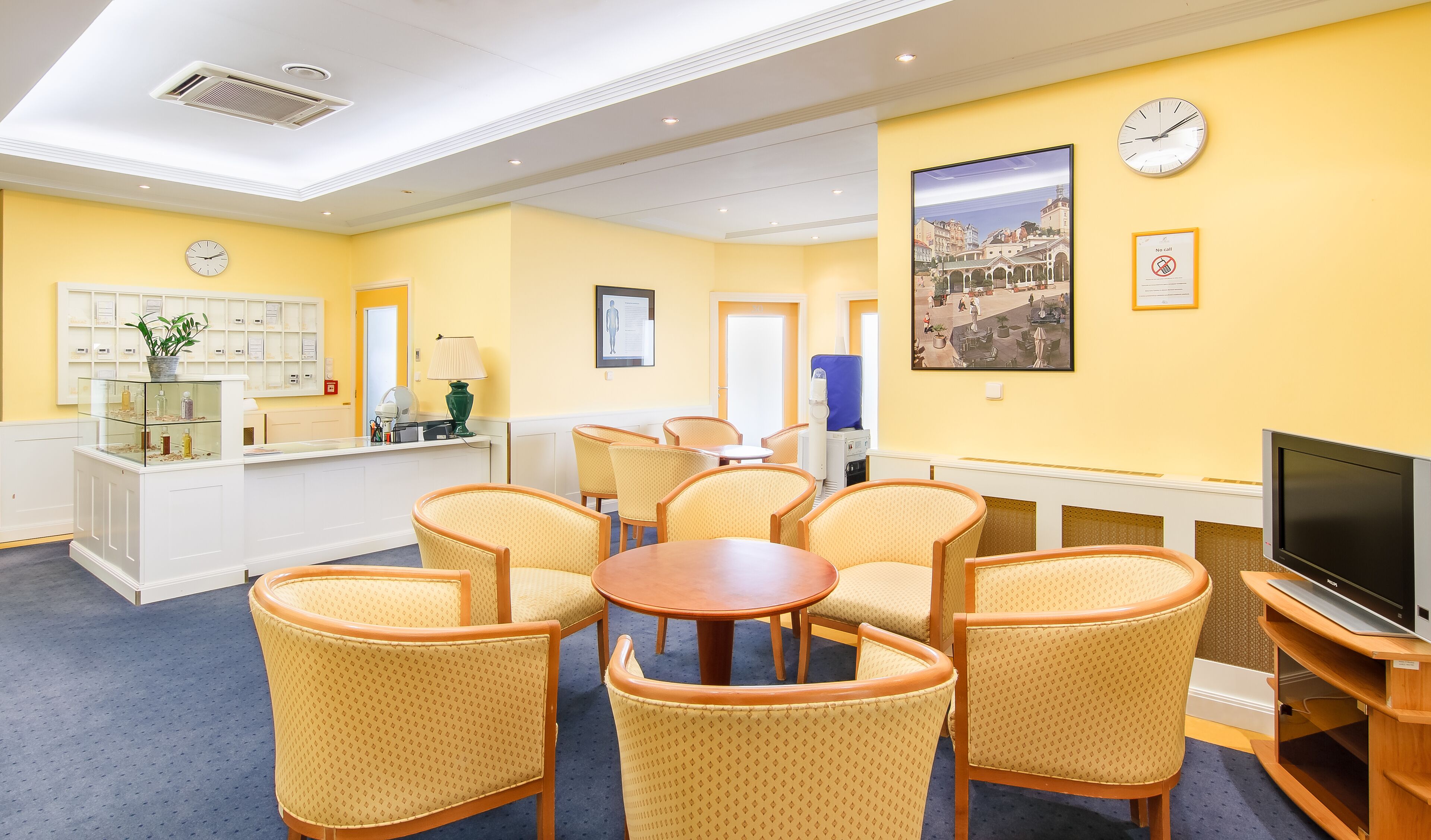 A hotel lobby with a reception desk, seating area with chairs, a television, and a clock on the wall.