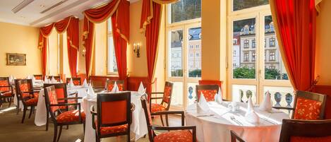 A dining area with tables set for service, red upholstered chairs, and large windows with red curtains.