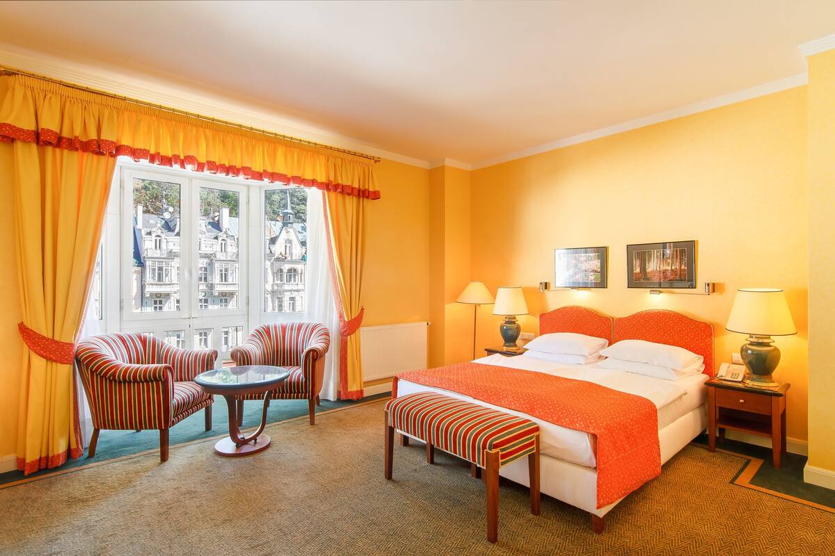 A hotel room with a large bed, two bedside lamps, a small table with a chair, a bench, and a window with curtains.