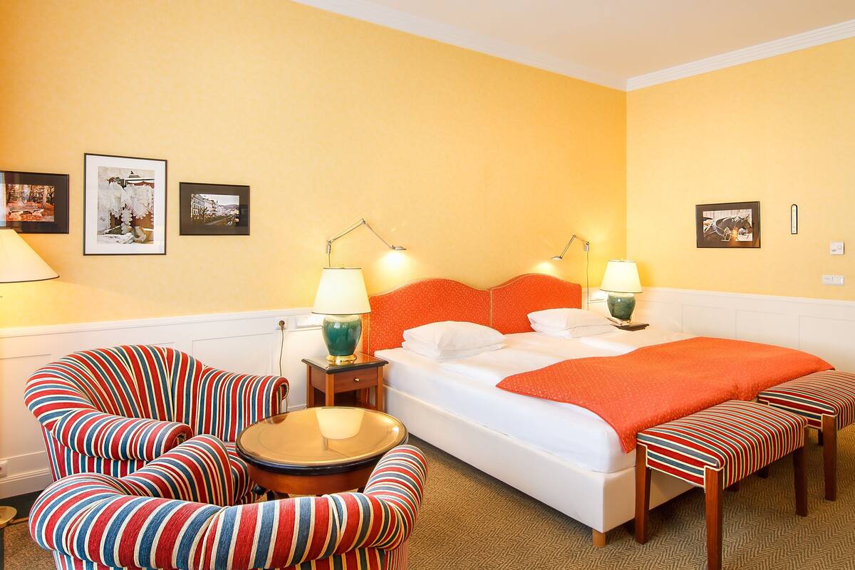 A hotel room with a bed, two bedside tables, a striped armchair, a bench, and a floor lamp.