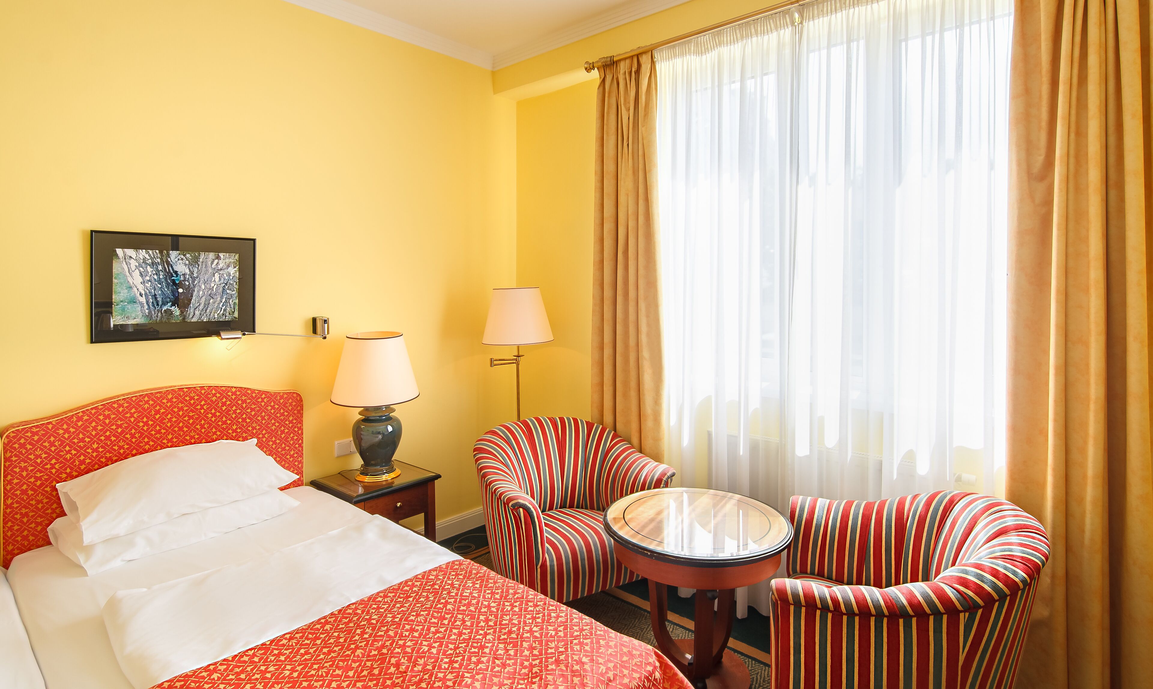 A hotel room with a bed, two striped armchairs, a small table, a lamp, and a painting on the wall.