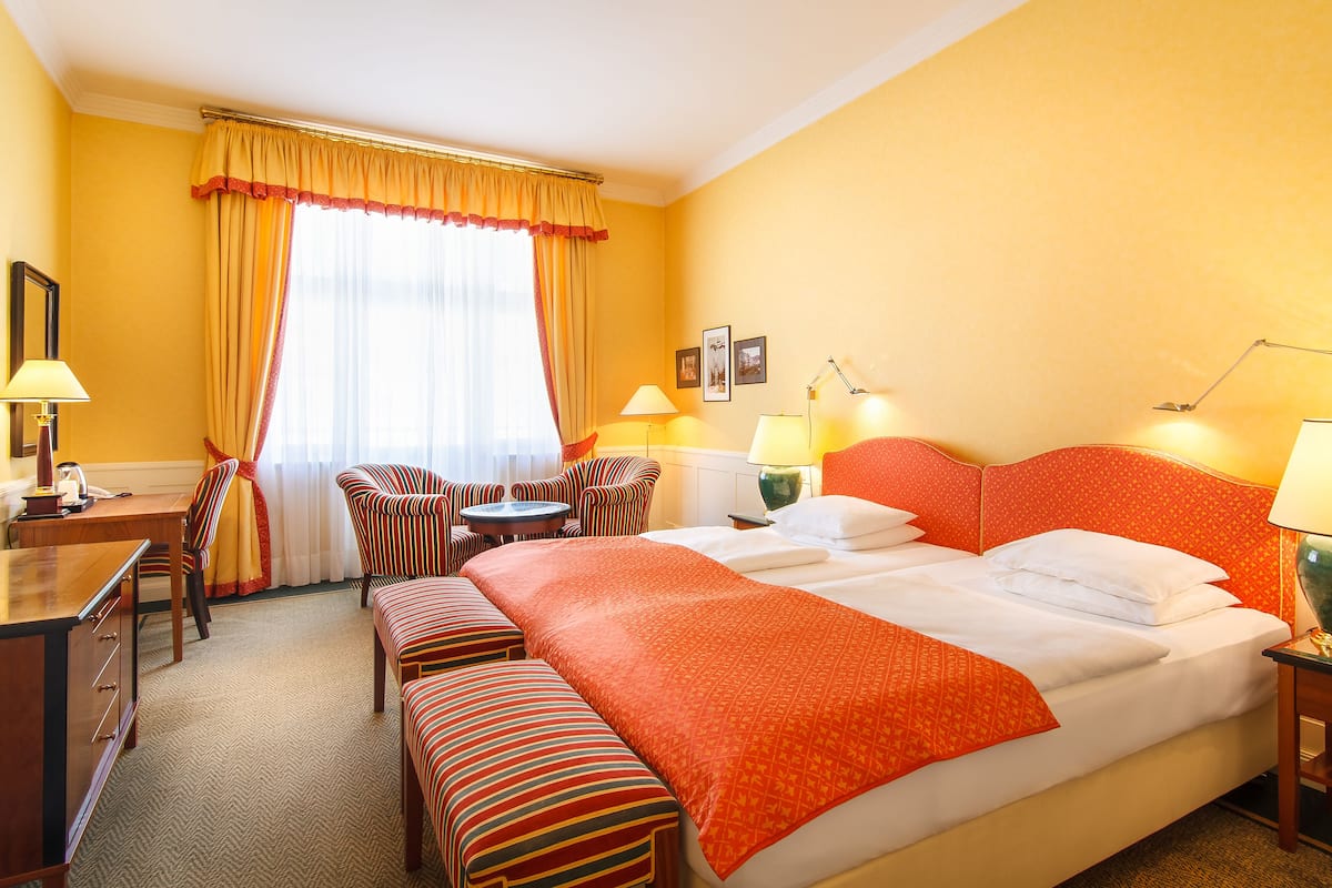 A hotel room with a large bed, a desk with a chair, a small table, and two ottomans.
