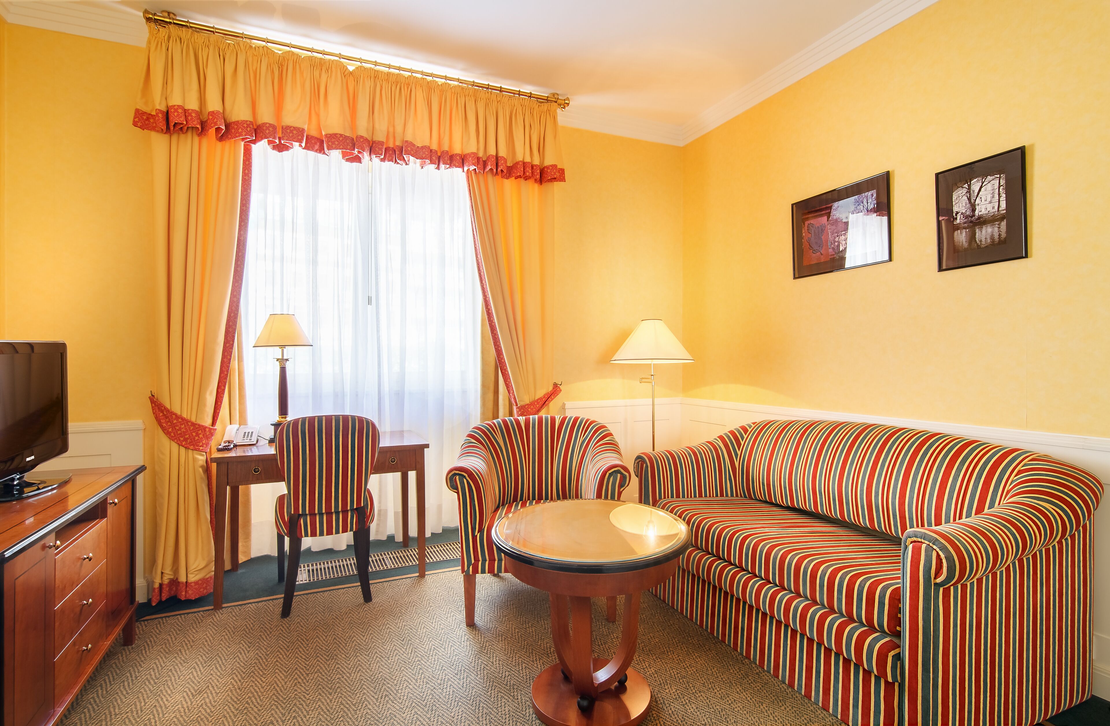 A hotel room with a striped sofa, a small round table, a desk with a lamp, a television, and two framed pictures on the wall.