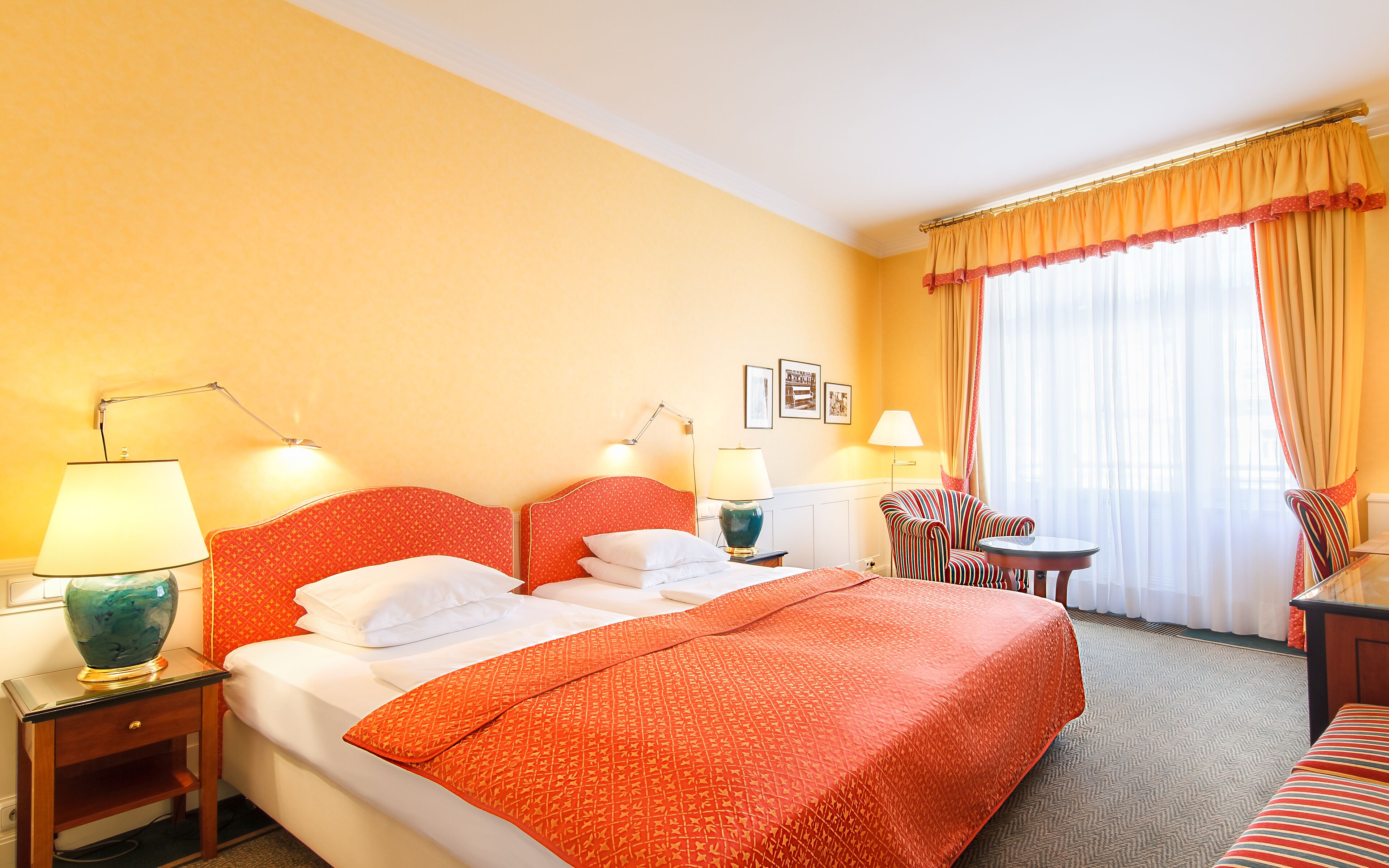 Habitación de hotel con una cama grande, dos mesitas de noche con lámparas, una mesa redonda pequeña, una silla y una ventana con cortinas.