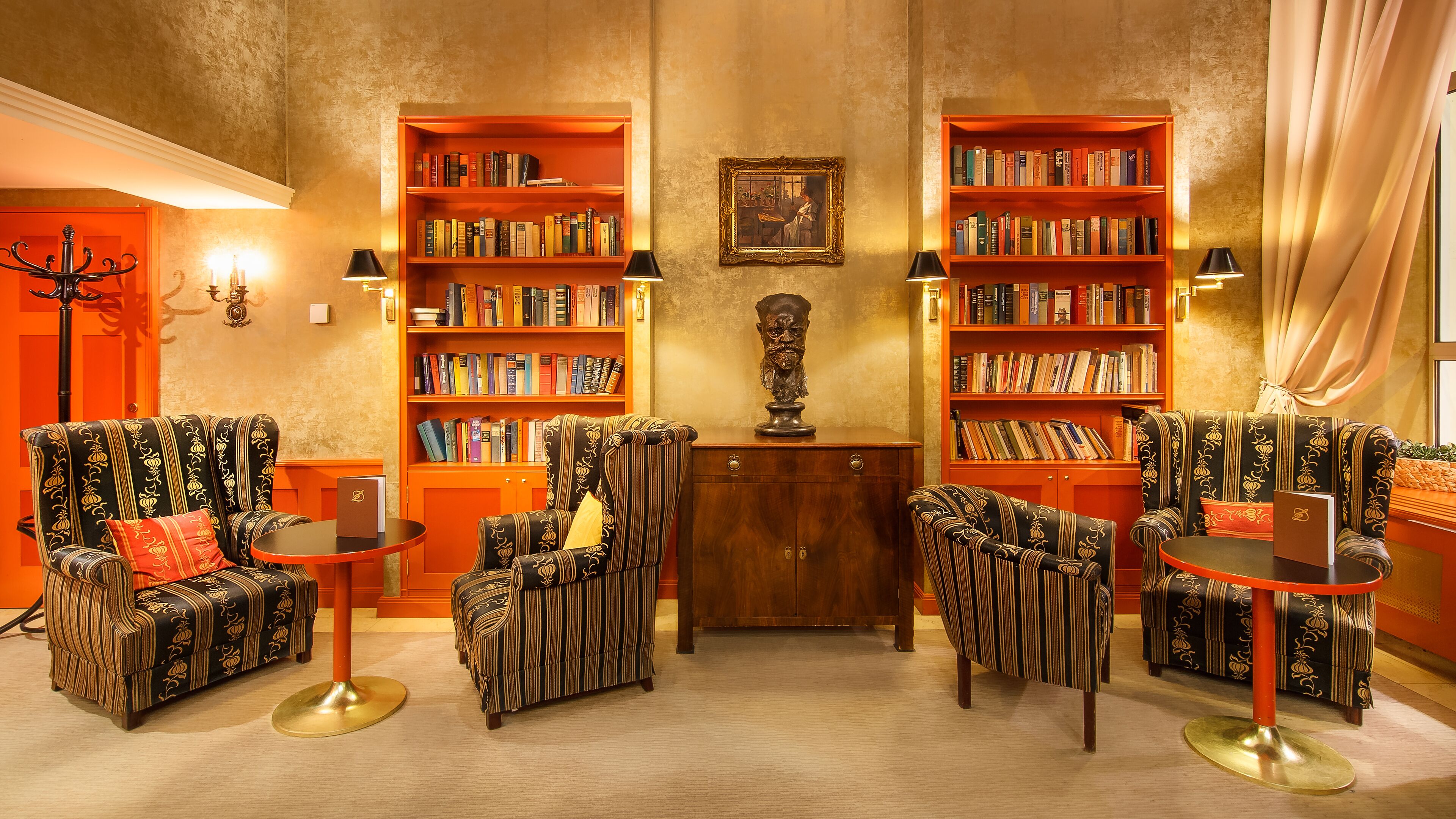A library with two large bookshelves filled with books, a bust sculpture, and striped armchairs with a red cushion.