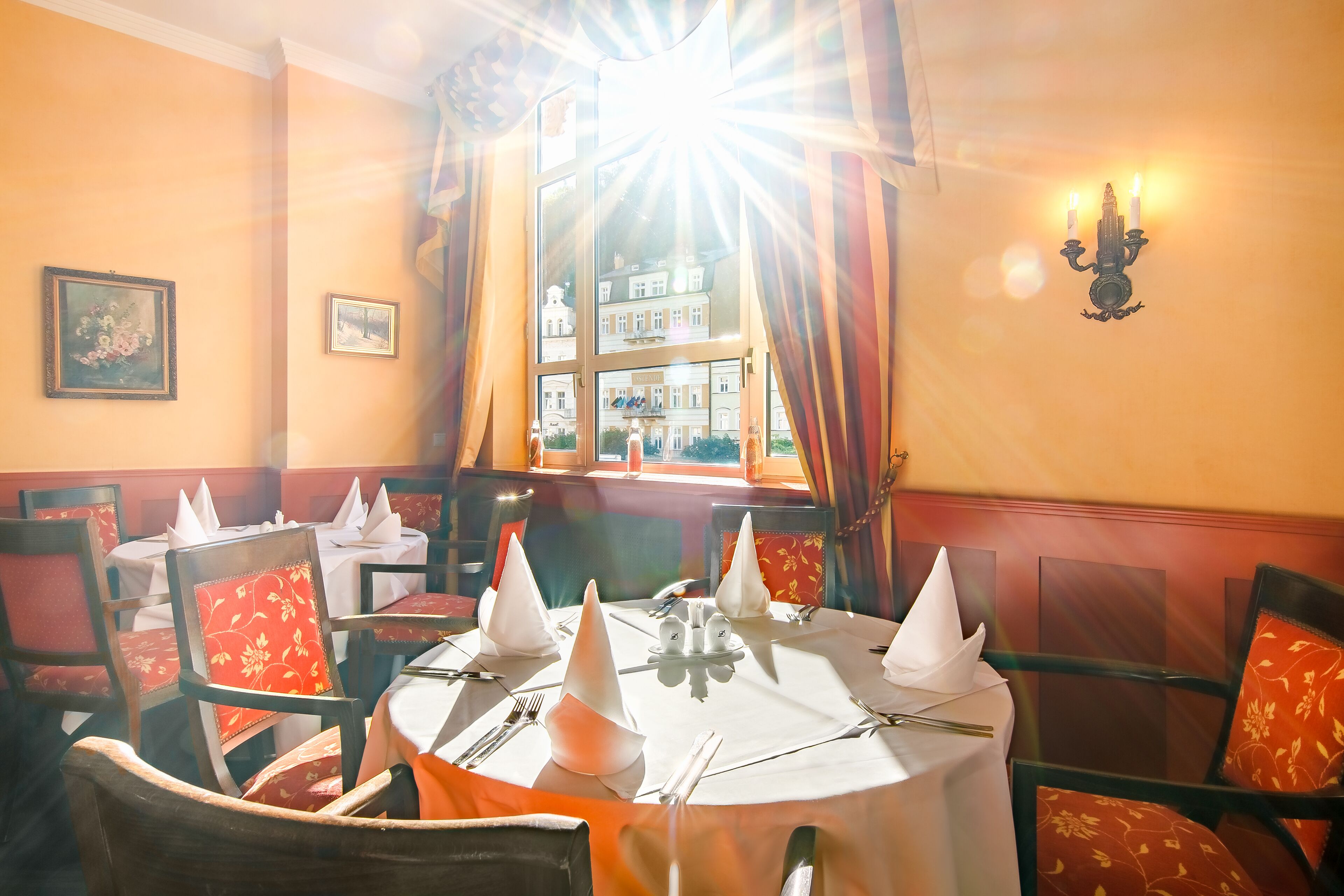 Comedor con mesas dispuestas para cuatro comensales, cada una con mantel blanco, servilletas y cubiertos. El salón cuenta con sillas tapizadas rojas con estampado floral.