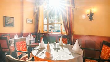 Comedor con mesas dispuestas para cuatro comensales, cada una con mantel blanco, servilletas y cubiertos. El salón cuenta con sillas tapizadas rojas con estampado floral.