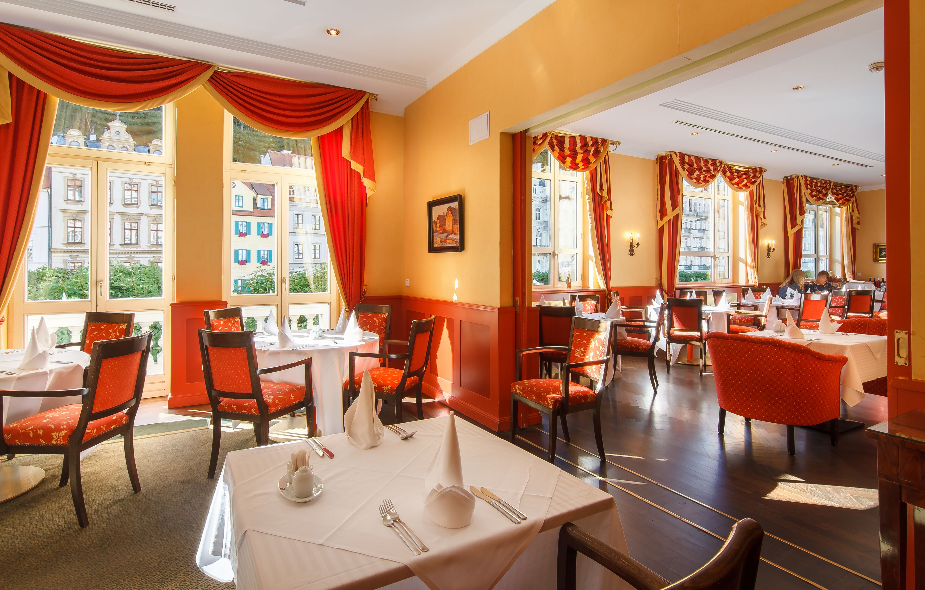 A restaurant with tables set for dining, red curtains, and large windows with a view of buildings.