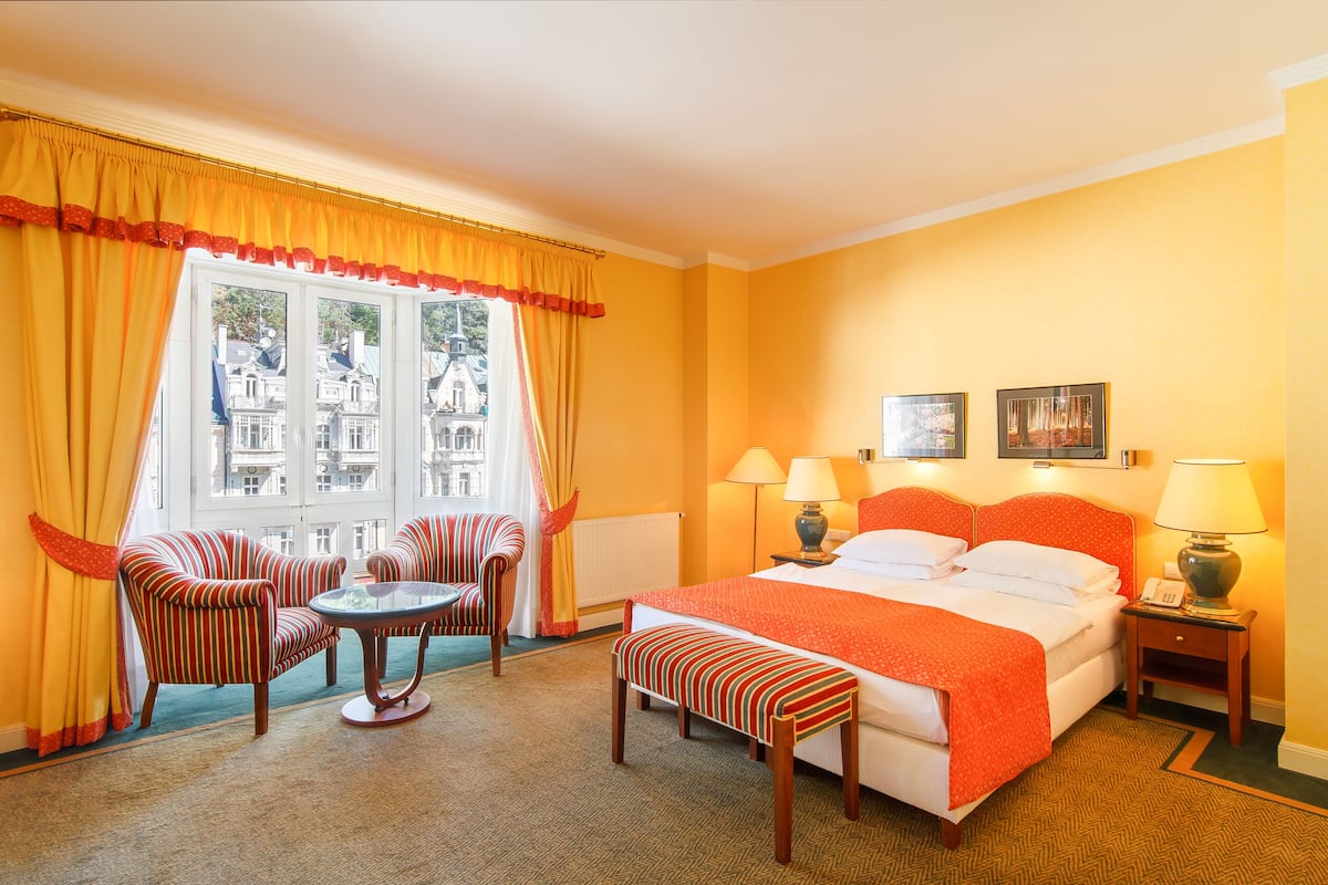 A hotel room with a large bed, two bedside lamps, a small table, two striped armchairs, a bench, and a window with curtains.