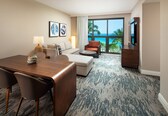 A hotel room with a wooden dining table, two sofas, a TV, and a large window with a view of the ocean.