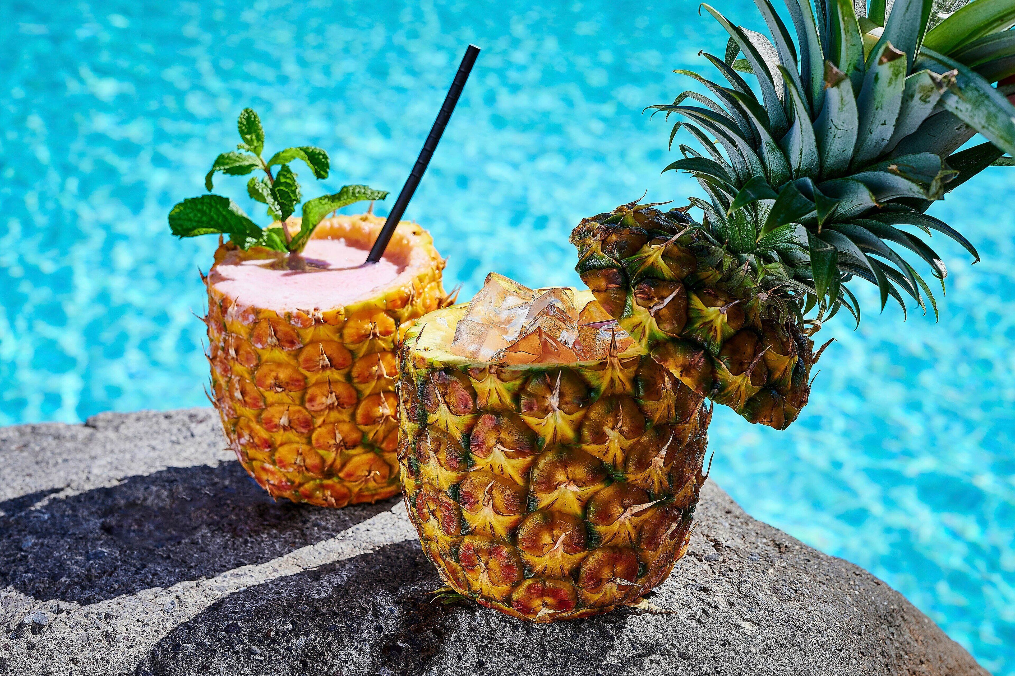 Deux boissons servies dans des demi-ananas, avec des pailles, posées sur une surface en pierre au bord d’une piscine.