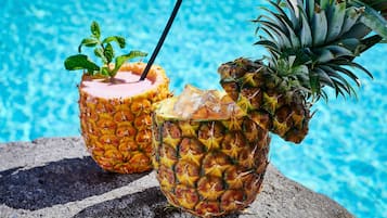 Deux boissons servies dans des demi-ananas, avec des pailles, posées sur une surface en pierre au bord d’une piscine.