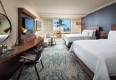 A hotel room with two beds, a desk with a chair, a small table, and a balcony with a view of palm trees.