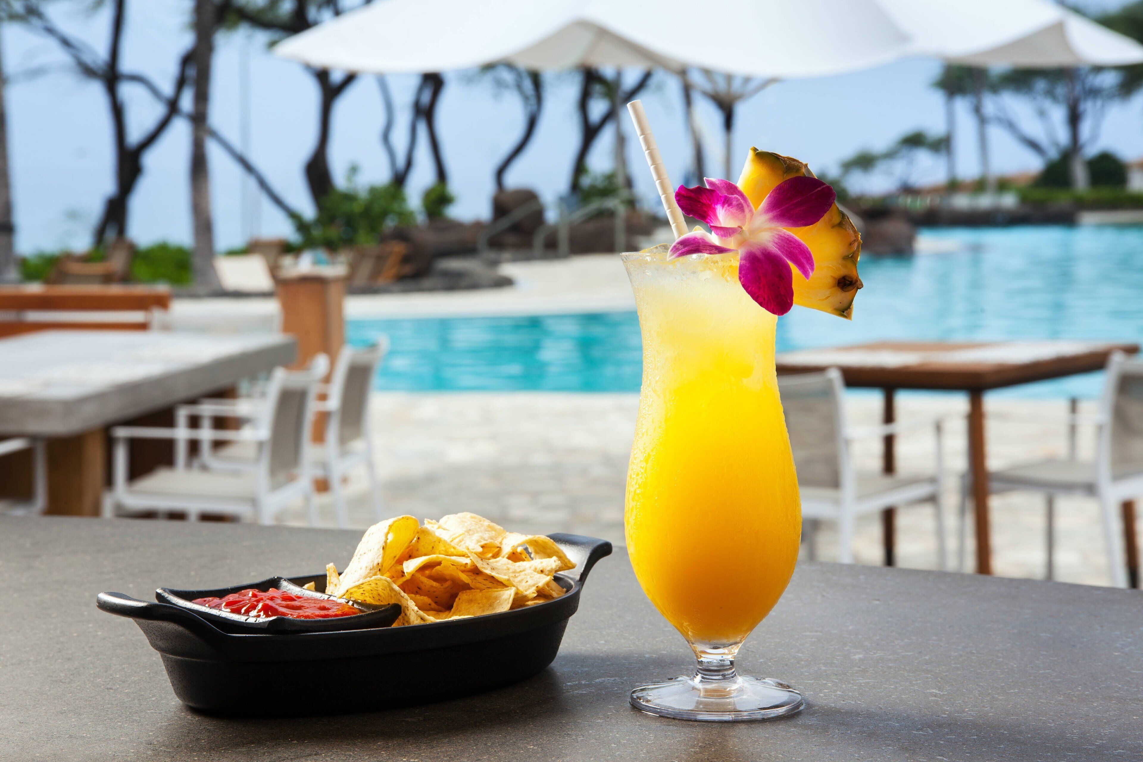 A table with a glass of orange juice and a bowl of chips by a poolside.