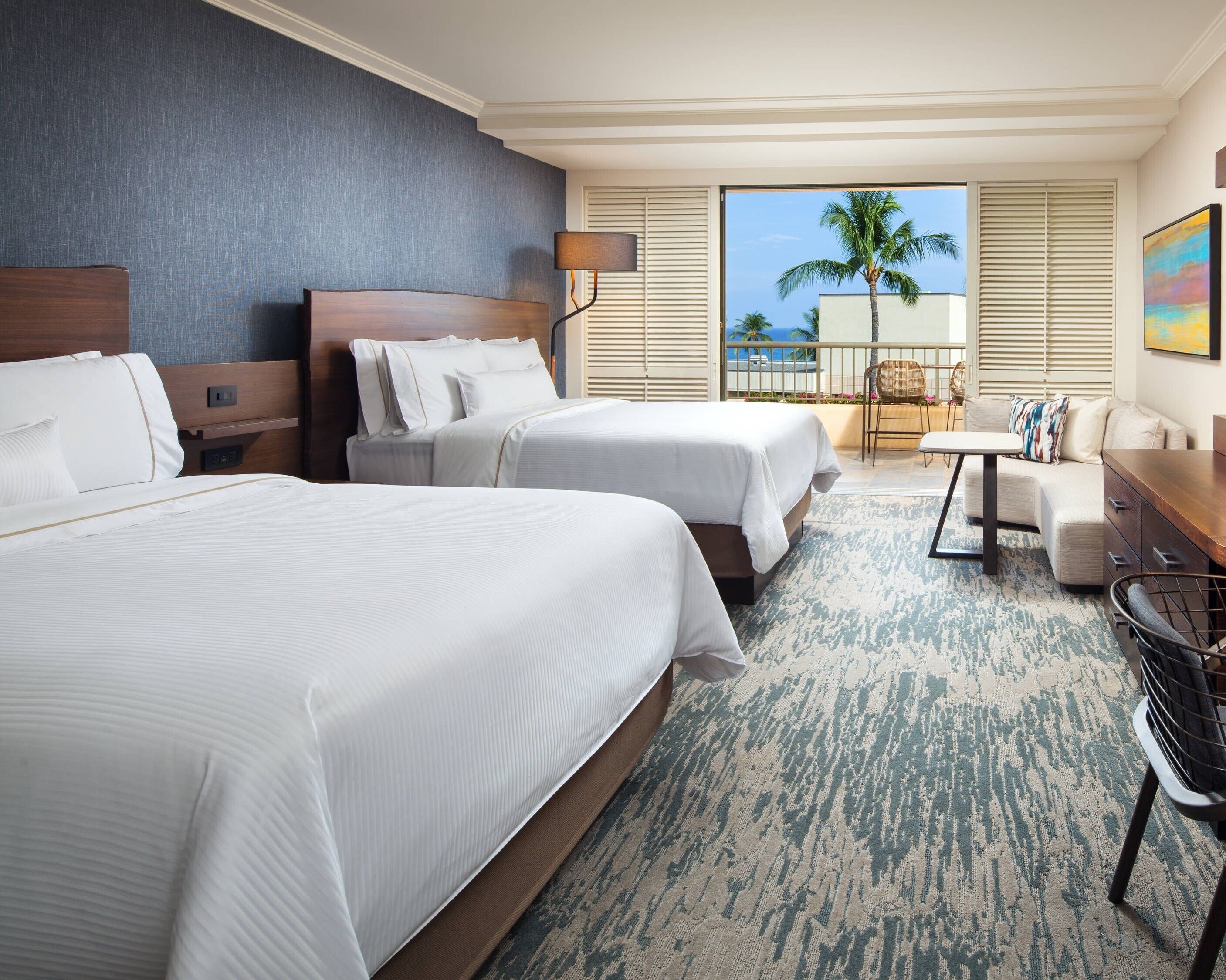 A hotel room with two beds, a sofa, a small table, and a view of palm trees and the ocean.