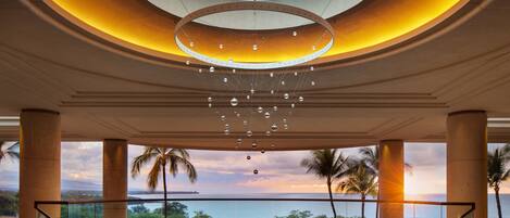 A spacious atrium with a central circular table, large pillars, and a view of the ocean at sunset.