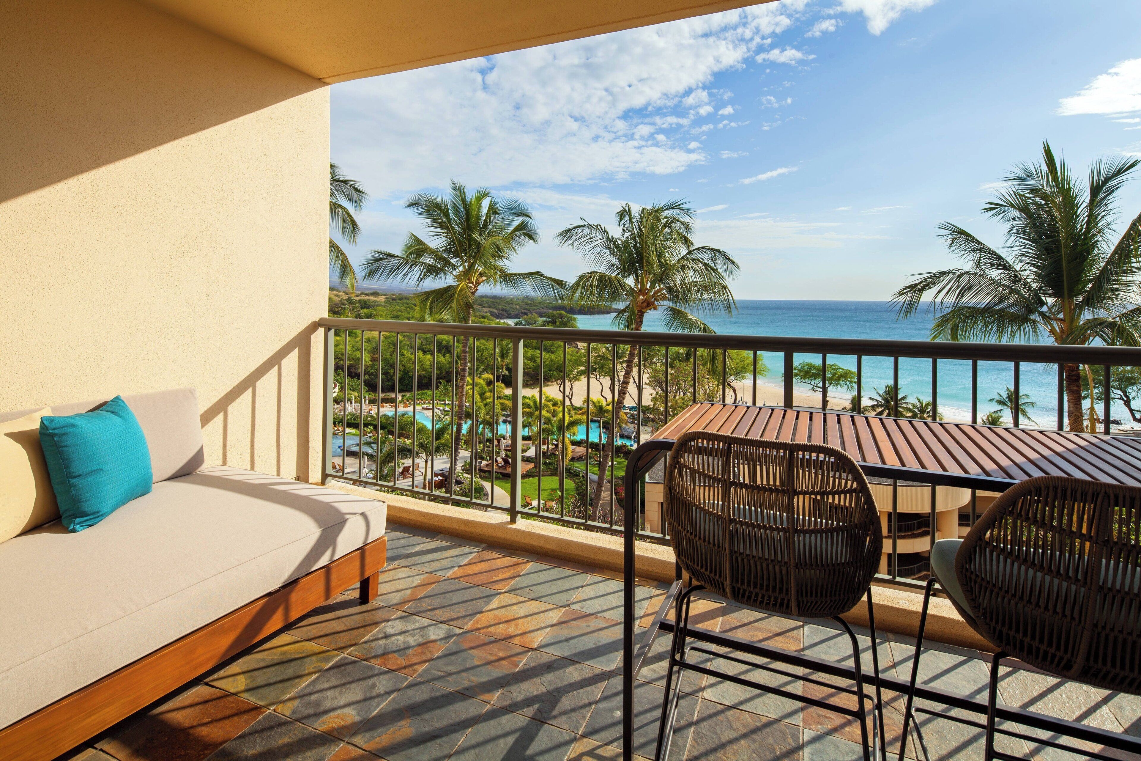 A balcony with a sofa, two wicker chairs, and a table with a view of the beach and ocean.