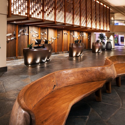 A spacious lobby with a wooden reception desk, large potted plants, and a stone floor.