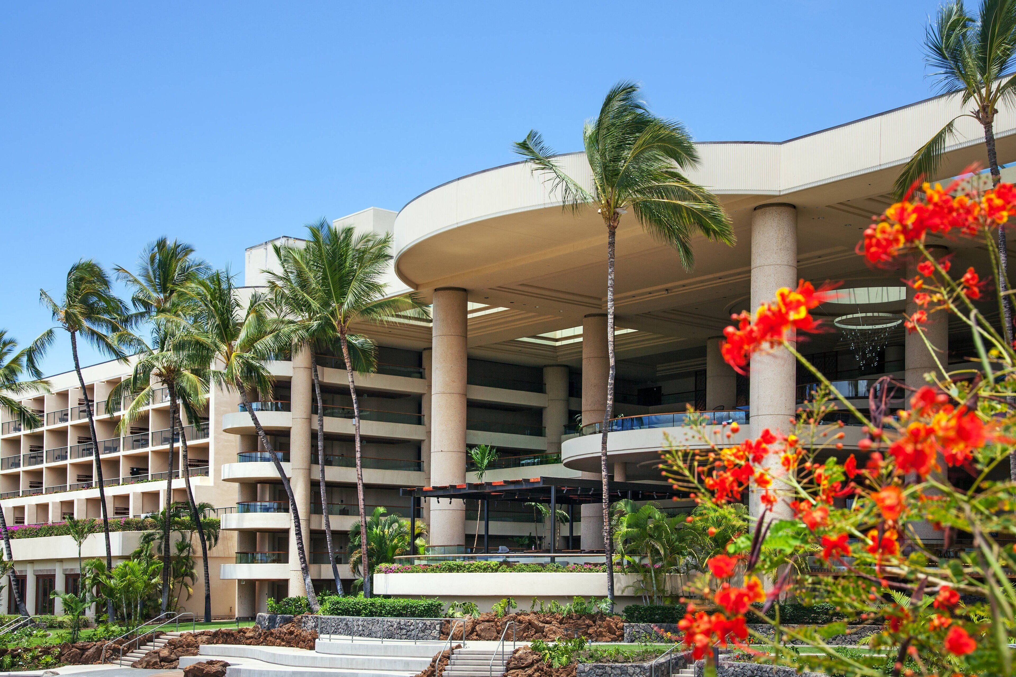 A multi-story hotel with a modern architectural design, featuring a central atrium with a curved roof, palm trees, and a landscaped garden area.