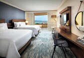 A hotel room with two beds, a desk, a chair, a large mirror, and a view of the sea.