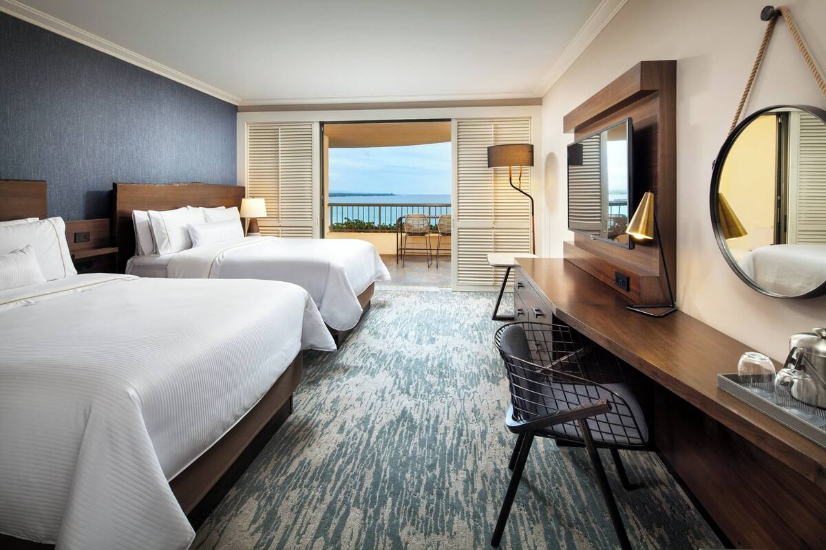 Une chambre d’hôtel avec deux lits, un bureau, une chaise, un grand miroir et une vue sur la mer.