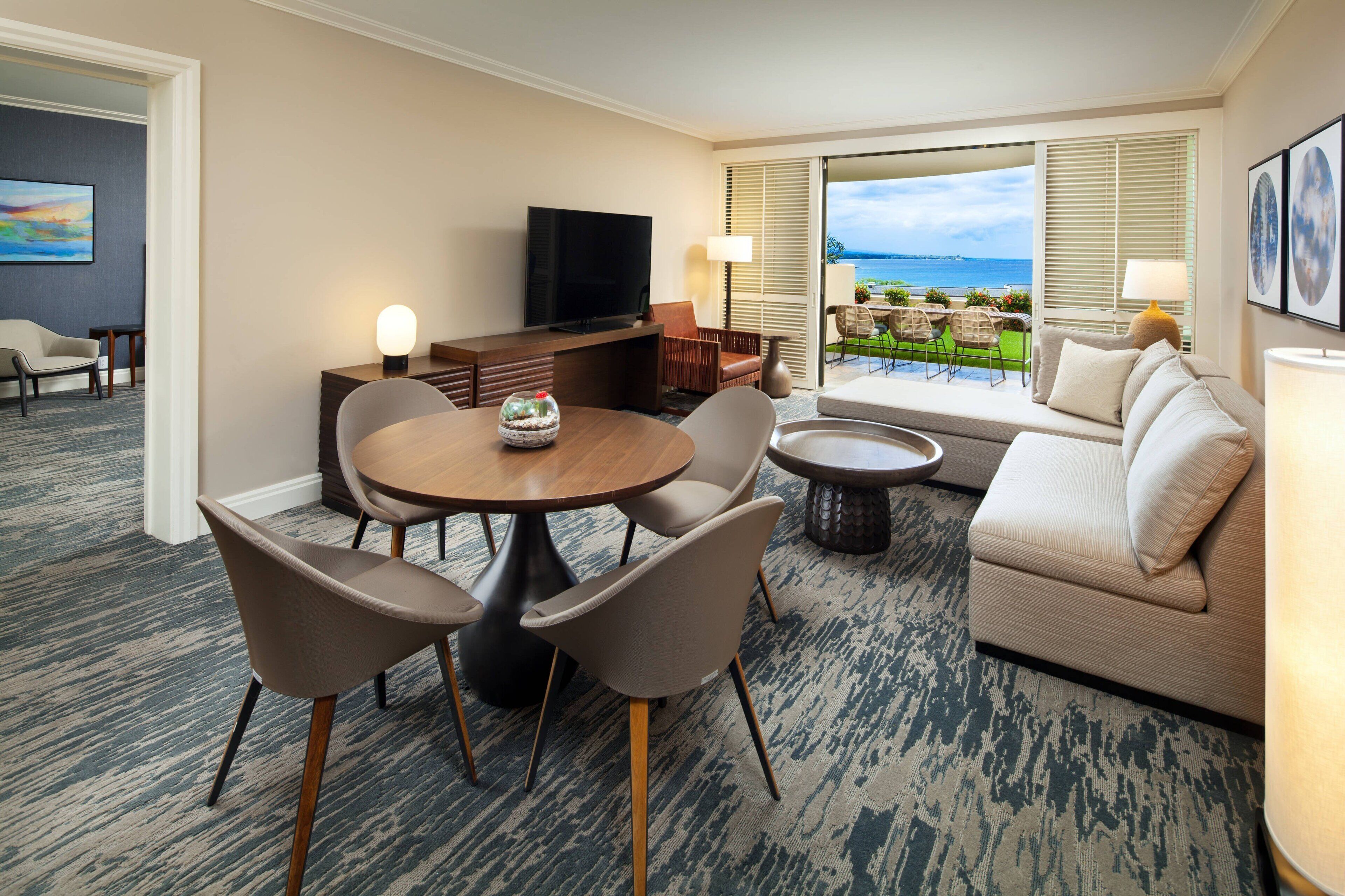 A hotel room with a dining area, a sofa, a TV, and a view of the ocean.