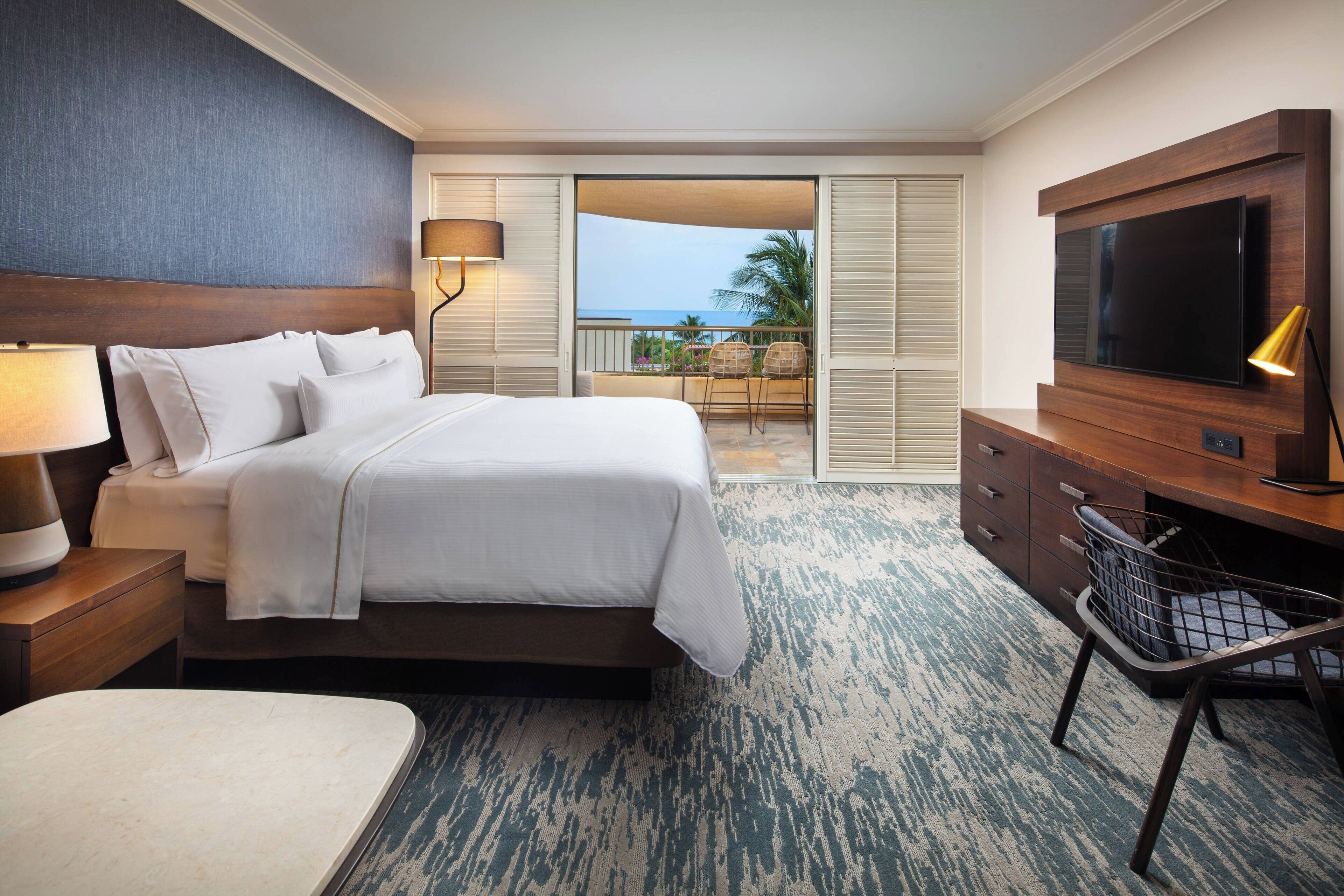 A hotel room with a bed, a desk, a TV, a lamp, and a view of the ocean.