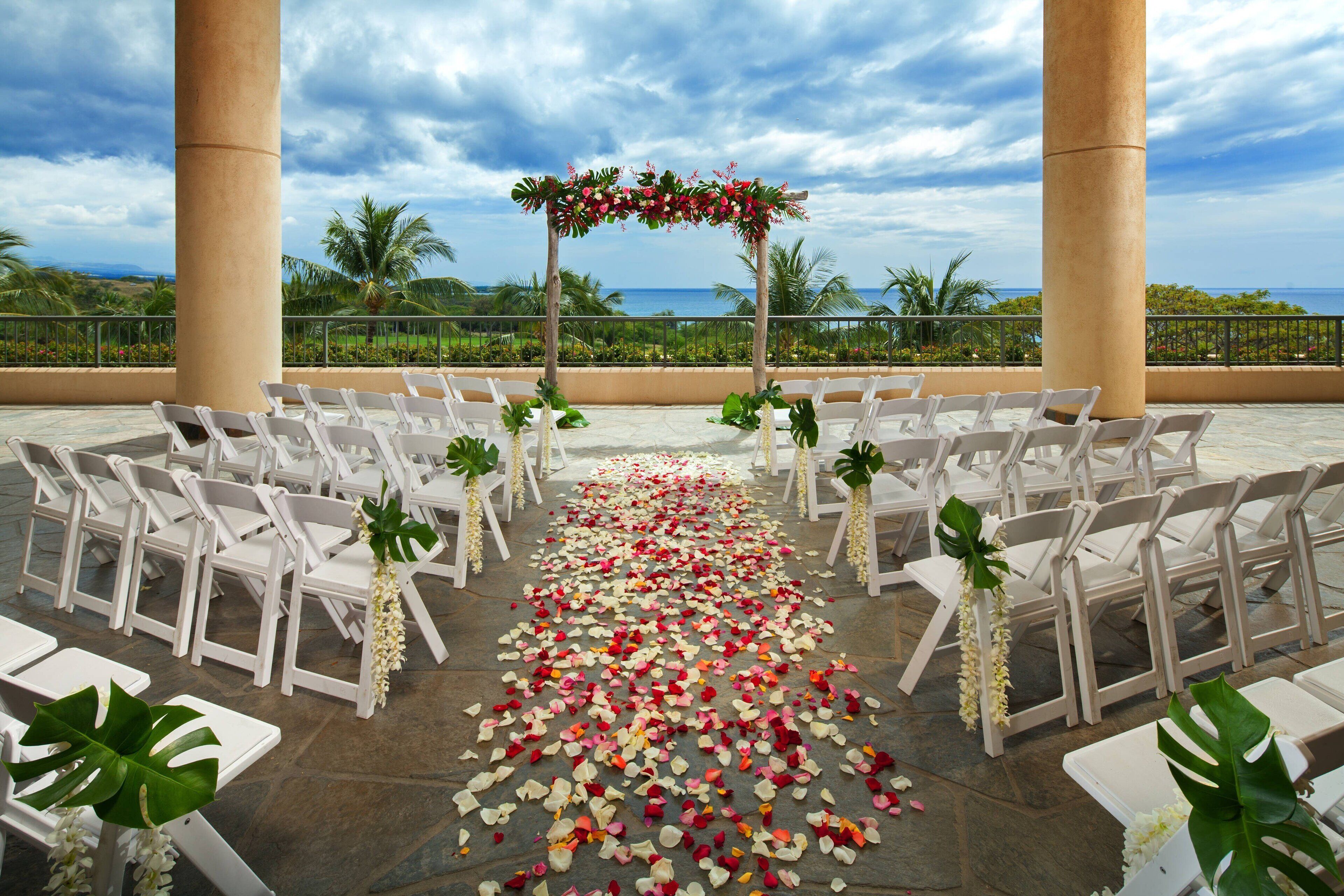An outdoor wedding aisle with white chairs and a floral arch, set against a backdrop of palm trees and the ocean.