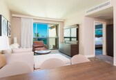 A hotel room with a large window, a sofa, a dining table, a TV, and a bed.