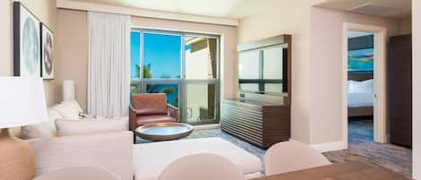 A hotel room with a large window, a sofa, a dining table, a TV, and a bed.