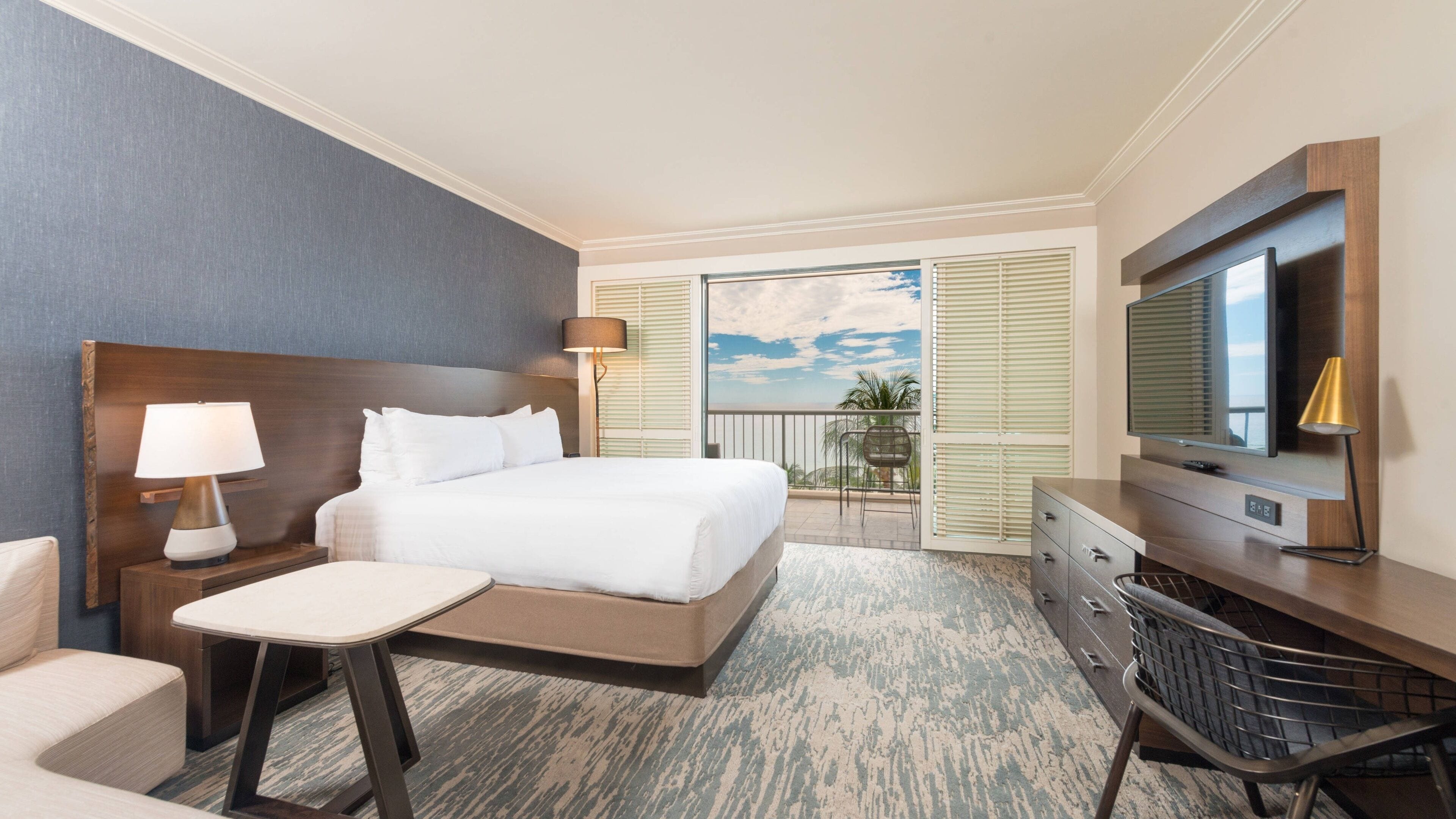 A hotel room with a large bed, a desk, a TV, and a balcony with a view.
