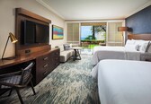 A hotel room with a large flat-screen TV, a desk with a lamp, two beds, a small table, and a view of the outdoors.