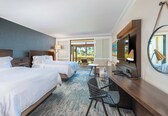A hotel room with two beds, a desk with a chair, a small table, a lamp, and a large mirror.
