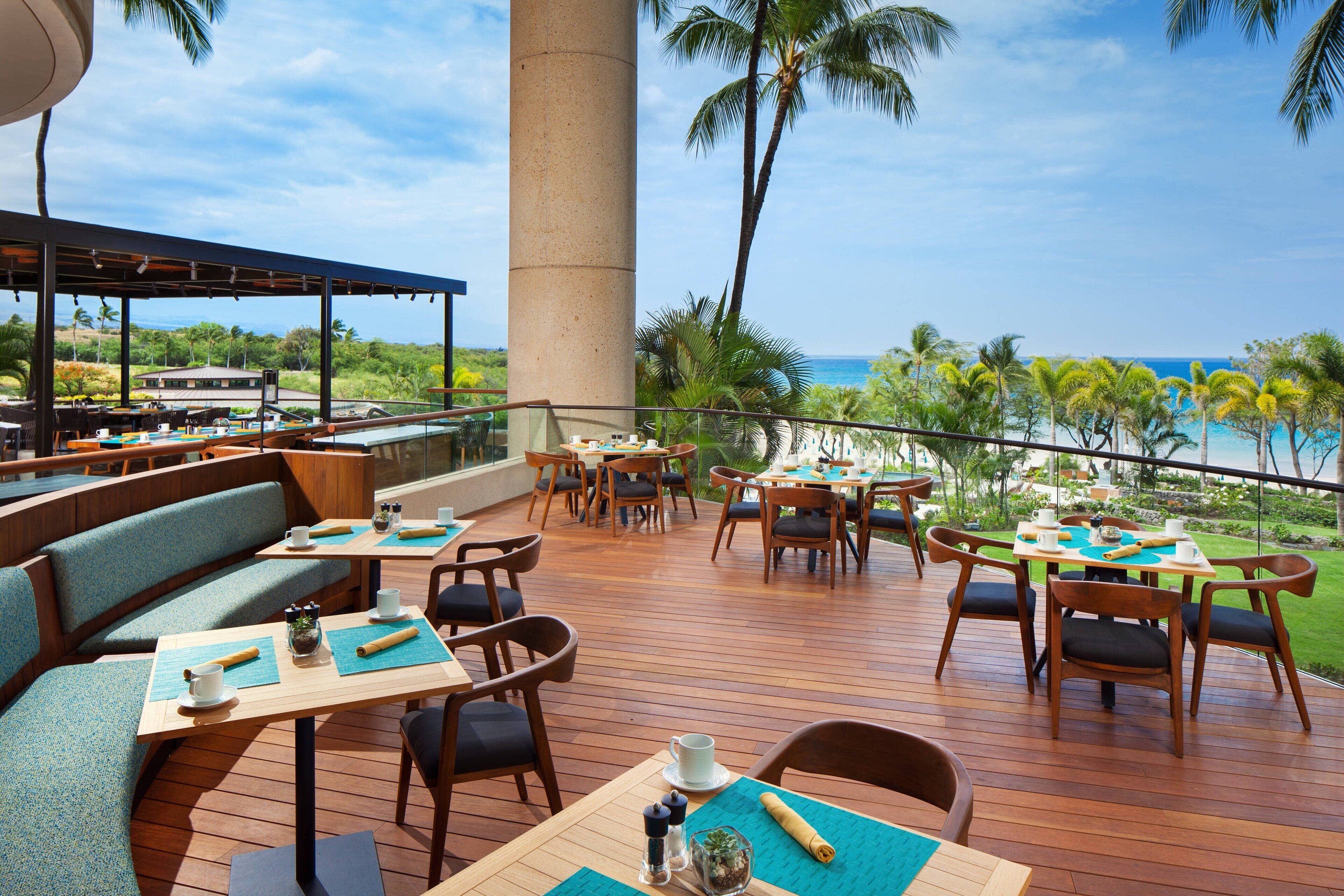 A restaurant deck with wooden tables and chairs, a large pillar, and a view of the ocean.